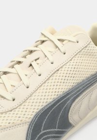 Nærbillede af beige sneakers med cremefarvede snørebånd, perforeret sidepanel og mørkegrå ruskinds Puma-logo på hvid baggrund.