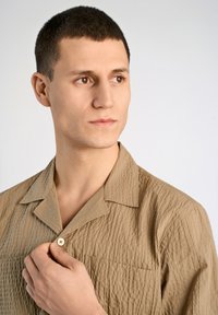 Camisa de manga corta texturizada en color beige con cuello tipo notch, un bolsillo en el pecho y cierre de botones. Tela suave con un patrón arrugado.