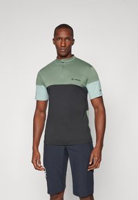 Camisa de manga corta con cremallera en bloques de color verde y gris oscuro. Presenta un cuello tipo mock y un logo en el pecho. Se combina con pantalones cortos navy.