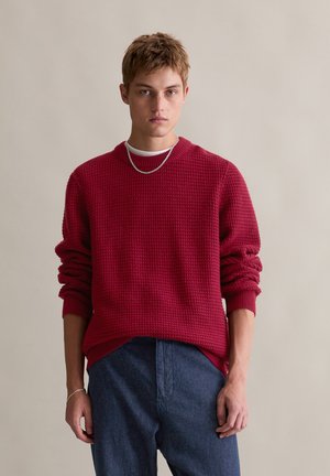 Roter strukturierter Pullover mit ribbed Rundhalsausschnitt und überschnittenen Schultern, kombiniert mit dunkelblauen Jeans. Eine silberne Kettenhalskette sorgt für das gewisse Etwas.