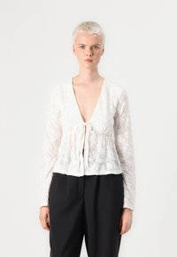 Weiße Spitzenbluse mit langen Ärmeln, tiefem V-Ausschnitt und bindbarem Frontdesign. Blumenmuster, weicher Stoff, kombiniert mit schwarzen Hose.