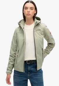 Lichtgroene softshell jack met kap, volledige rits en geborduurd logo. Gecombineerd met een crèmekleurige trui en blauwe denim. Bevat ritszakken.