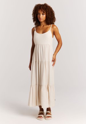 Vrouw met krullend haar, gekleed in een mouwloze, gelaagde beige maxi-jurk en beige sandalen, staand tegen een effen lichte achtergrond.