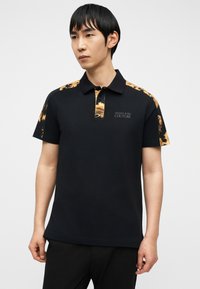 Άνδρας που φοράει μαύρο πόλο με χρυσό μπαρόκ σχέδιο στους ώμους και το γιακά, στέκεται μπροστά σε απλό φόντο.