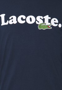 T-shirt en coton bleu marine avec un grand texte blanc "Lacoste" et un logo crocodile brodé vert avec une bouche rouge. Texture douce.