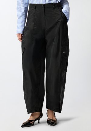 Osoba oblečená v čiernych širokých nohaviciach s vysokým pásom s gombíkovými cargo vreckami, v kombinácii s modrou pruhovanou košeľou a čiernymi špicatými podpätkami.