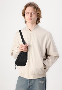 Beige bomberjakke med lynlås foran, ribbestrikkede manchetter og kant; parret med en lille sort crossbody taske med lynlåslukning og strop.