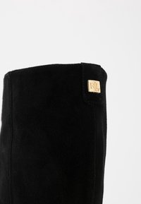 Černé semišové kozačky do kolen s hladkou texturou, s kultivovaným zlatým obdélníkovým logem na boku. Jednoduchý, elegantní design.