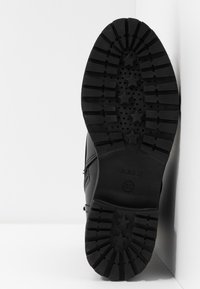 Semelle de bottine noire avec des crampons en forme d'étoile et des rainures profondes, reposant verticalement contre un mur blanc d'angle.