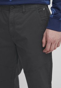 Pantaloni negri cu fața netedă, cu curea și buzunare laterale. Prezintă o textură fină și cusături vizibile pe genunchi.