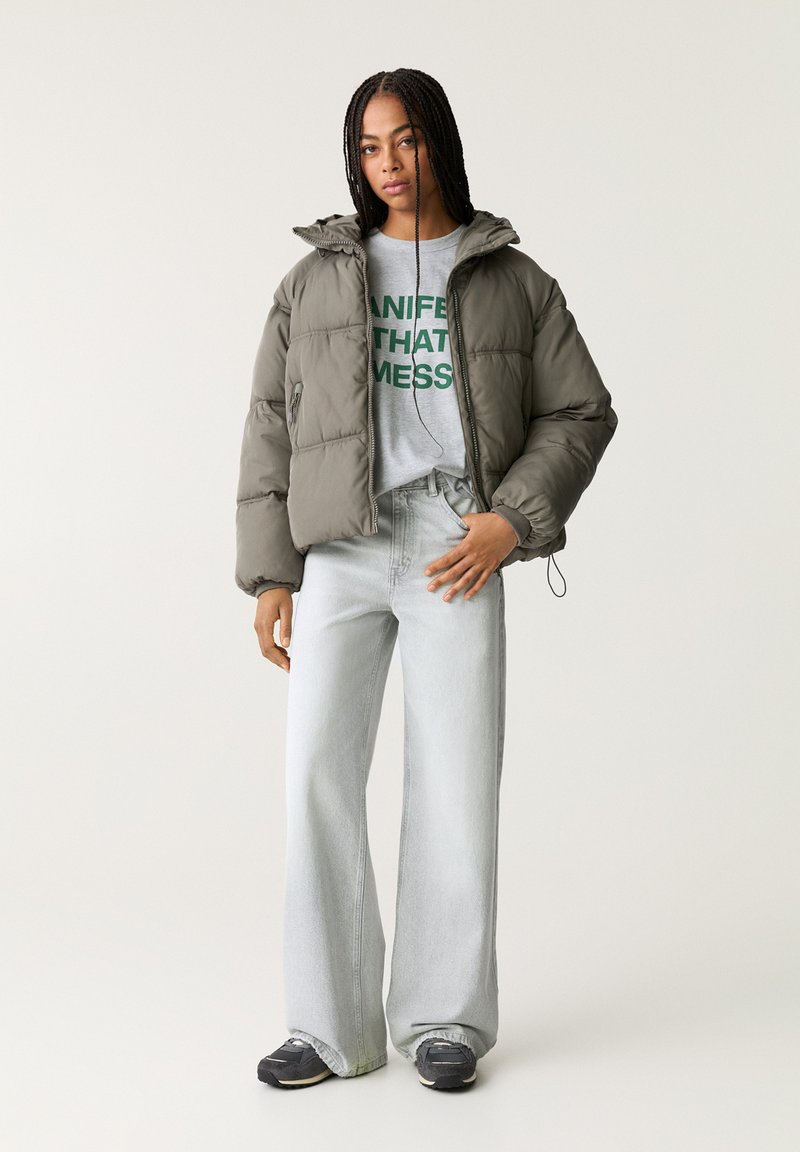 Giacca a vento in verde oliva scuro con cappuccio, chiusura a zip e tasche laterali; abbinata a una maglietta grigia con testo verde e jeans azzurri chiari.
