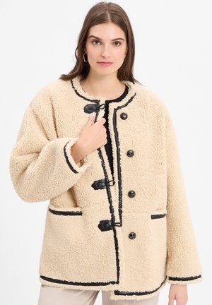 Beige fleecespecial met zwarte accenten, knopen aan de voorkant, oversized pasvorm en opgestikte zakken. Heeft een lussluiting en een zachte textuur.