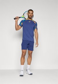 Sinine sportlik t-särk mitmevärviliste mustriga õlgadega, koos siniste lühikeste pükstega ja valgete spordijalatsitega. Hoides tennisereketit.