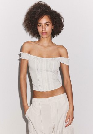 Kobieta o kręconych włosach, ubrana w białą bluzkę typu crop top z odkrytymi ramionami oraz jasne spodnie, stojąca na tle jednolitego białego tła.