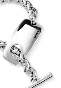 Bracelet en chaîne argentée avec une grande étiquette rectangulaire au surface polie et logo gravé, mettant en avant un mélange de maillons et de quincaillerie.