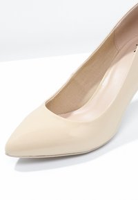 Beige patent-nahkainen avokärkikorkokenkä, jossa on terävä kärki ja keskipitkä korko. Sisäpuolella on sileä, kontrastoiva beigevuori.