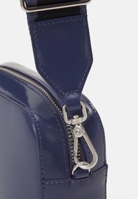 Borsa in pelle vernice blu navy con angolo, dotata di zip e accessori in metallo argentato, caratterizzata da una texture liscia e dettagli di cucitura rinforzati.