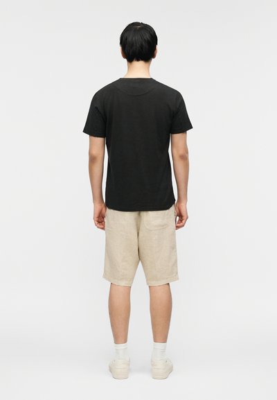 Μαύρο βαμβακερό t-shirt με κοντά μανίκια, σε συνδυασμό με μπεζ λινά σορτς. Το σύνολο ολοκληρώνεται με λευκές κάλτσες και μπεζ αθλητικά παπούτσια.