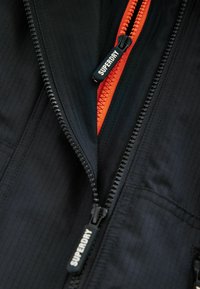 Giacca nera con un tessuto texture, dotata di una cerniera nera e una cerniera interna arancione. Il marchio "SUPERDRY" è visibile sul cursore della cerniera.