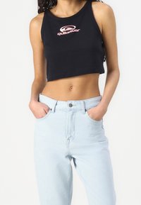 Kobieta w czarnym crop topie Quiksilver i jasnoniebieskich, wysokich spodniach dżinsowych, z rękami w kieszeniach na białym tle.