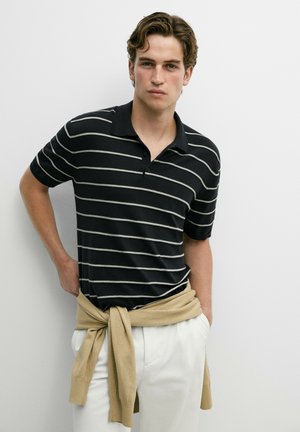 Giovane uomo che indossa una polo a righe bianche e nere e pantaloni bianchi con un maglione beige legato intorno alla vita, in piedi contro un muro bianco.