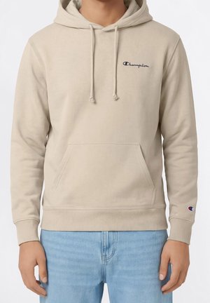 Mann trägt beigen Champion Hoodie mit Fronttasche und hellblauen Jeans, gezeigt von den Schultern bis zur Hüfte vor weißem Hintergrund.