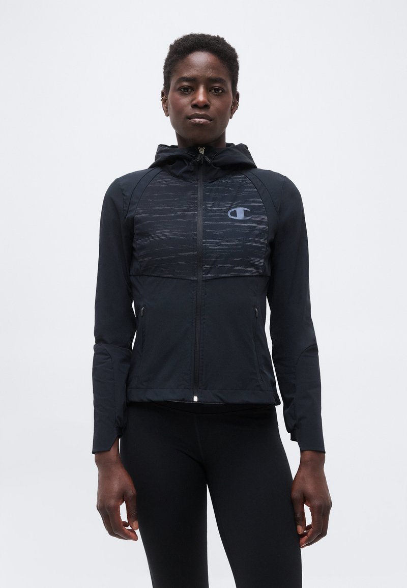 Νέο άτομο με μαύρο δέρμα, που φοράει μαύρο φούτερ με φερμουάρ και κουκούλα, με λογότυπο στο στήθος, στέκεται μπροστά σε απλό ανοιχτό φόντο.