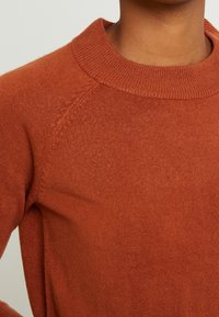 Maglione lavorato a maglia di colore arancio ruggine con scollatura rotonda a costine e maniche raglan, caratterizzato da una texture morbida e senza motivi o decorazioni visibili.