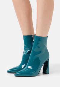 Botines de cuero charol color teal con punta afilada, cierre de cremallera y tacón grueso alto. Textura suave y diseño elegante.