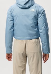 Niebieska krótka kurtka z kapturem, wykonana z gładkiego materiału, z białym logo na górnej części pleców oraz elastycznymi mankietami. W zestawie z beżowymi spodniami.