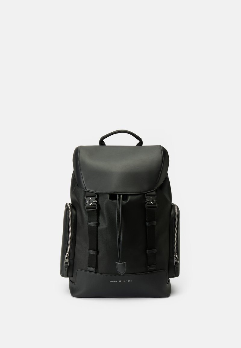 Sac à dos noir en simili cuir avec une fermeture à rabat, des sangles avant, des poches zippées latérales et une poignée supérieure. Texture lisse et design minimaliste.