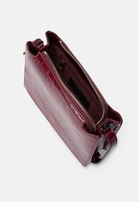 Borsa a tracolla in pelle bordeaux con finitura lucida, chiusura con zip e tracolla singola. L'interno presenta una fodera scura e una tasca.