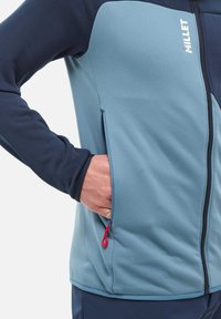 Kurtka softshell w jasnoniebieskim i granatowym kolorze. Posiada podniesiony kołnierz, boczne kieszenie zapinane na zamek z czerwonymi uchwytami oraz gładką teksturę.