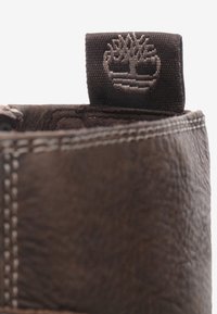 Hnědé kožené boty s texturovaným povrchem, zdobené tmavohnědým látkovým štítkem s vyraženým logem stromu na límci.