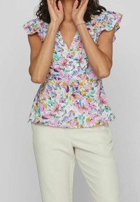 Femme portant un chemisier à fleurs pastel avec col en V et manches à volants, et un pantalon de couleur claire, les mains levées près du visage, sur un fond uni.