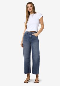 Νεαρή γυναίκα με μακριά ίσια μαλλιά, φορώντας λευκή κοντομάνικη μπλούζα, ψηλόμεσα φαρδιά τζιν σε μπλε απόχρωση και καφέ ανοιχτό παπούτσια με τακούνι, στέκεται με το χέρι στην τσέπη.