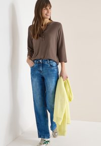Femme souriante, portant un haut marron à manches longues, un jean bleu ample, des baskets vertes et blanches, tenant un sweat-shirt jaune plié.