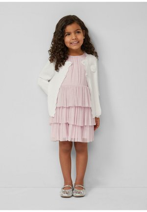 Ragazza giovane con capelli ricci che indossa un cardigan bianco, un vestito rosa chiaro a strati e scarpe Mary Jane argentate, in piedi davanti a uno sfondo semplice.