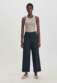 Vaaleanruskea hihaton paita, jossa on sinisiä yksityiskohtia, yhdistetty tummansinisisiin korkea vyötäröisiin leveälahkeisiin housuihin. Pehmeä materiaali ja rento istuvuus.