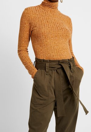 Femme portant un col roulé orange côtelé rentré dans un pantalon taille haute vert olive ceinturé avec de grandes poches, mains dans les poches.