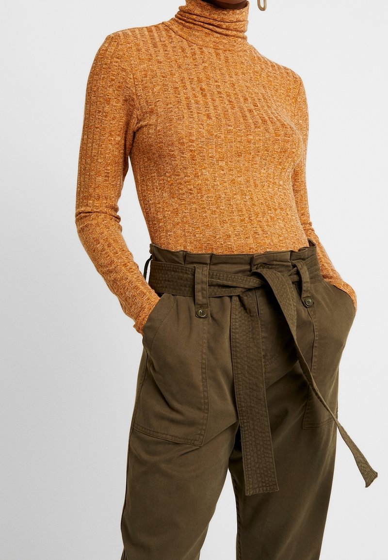 Femme portant un col roulé orange côtelé rentré dans un pantalon taille haute vert olive ceinturé avec de grandes poches, mains dans les poches.