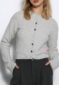 Jasnoszary dzianinowy kardigan z okrągłym dekoltem, długimi rękawami i skróconym dołem. Posiada ciemne guziki oraz małą różową naszywkę z logo.