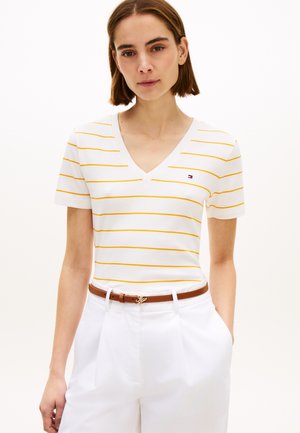 Femeie purtând tricou alb cu gât în V și mâneci scurte, cu dungi galbene subțiri, pantaloni albi și curea maro, stând cu mâinile în buzunare.