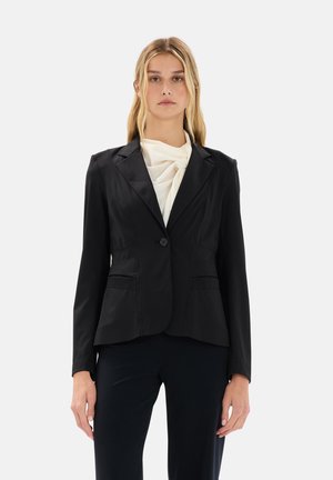 Blazer nero su misura con un solo bottone, due tasche frontali e revers a punta. Indossato sopra una blusa bianca a trama. Finitura in tessuto liscia.