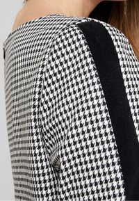 Hedvábná černobílá tkanina ve vzoru houndstooth, vršek s pevným černým panelem podél ramene. Hladká textura s kulatým výstřihem.