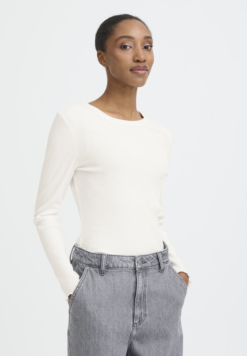 Sieviete ar rokām kabatās, ģērbusies baltā ribotā garām piedurknēm kreklā un augstās jostasvietas pelēkās džinsās, stāv pret vienkāršu baltu fonu.