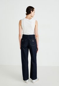 Jeans in denim scuro a vita alta con gamba dritta, dotati di due tasche posteriori, cuciture gialle e orlo risvoltato. Indossati con un top senza maniche bianco.