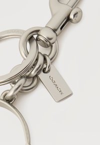 Μπρελόκ από ασημένιο μέταλλο με πολλαπλούς κρίκους και ορθογώνια ετικέτα χαραγμένη με τη λέξη «COACH», εμφανής αγκράφα πρόσδεσης σε λευκό φόντο.