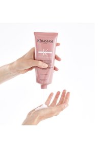 Hænder, der doserer hvid creme fra en lyserød Kérastase Chroma Absolu balsamtube på en åben håndflade mod en hvid baggrund.