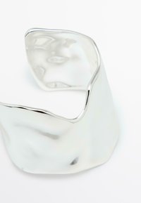 Metallarmband i form av en cuff med en polerad, vågig yta. Den har en oregelbunden form som reflekterar ljus med en glänsande silvrig finish.